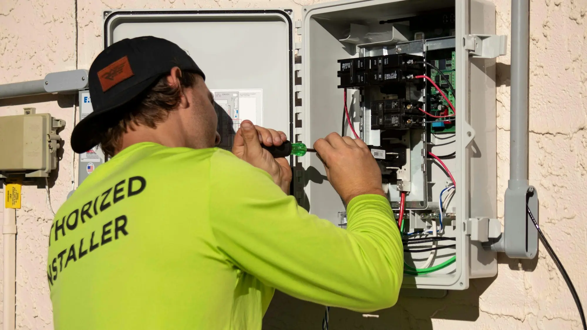 Electrician in a yellow work shirt using a screwdriver on an open outdoor electrical panel on a house wall
