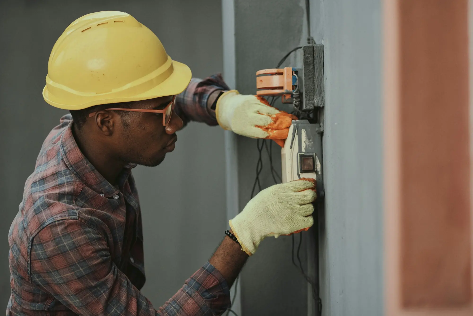 Electrician in a yellow hard hat and work gloves repairing a wall-mounted electrical switch