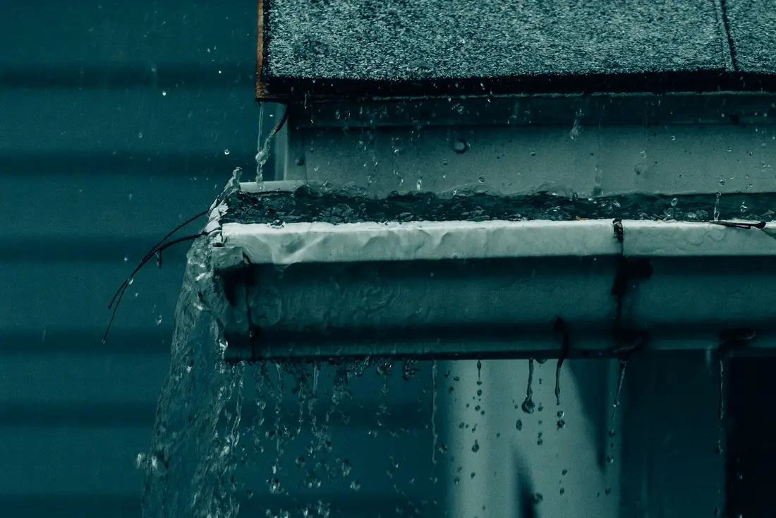 Rainwater spilling over the edge of a clogged white gutter beneath a gray shingle roof on a house