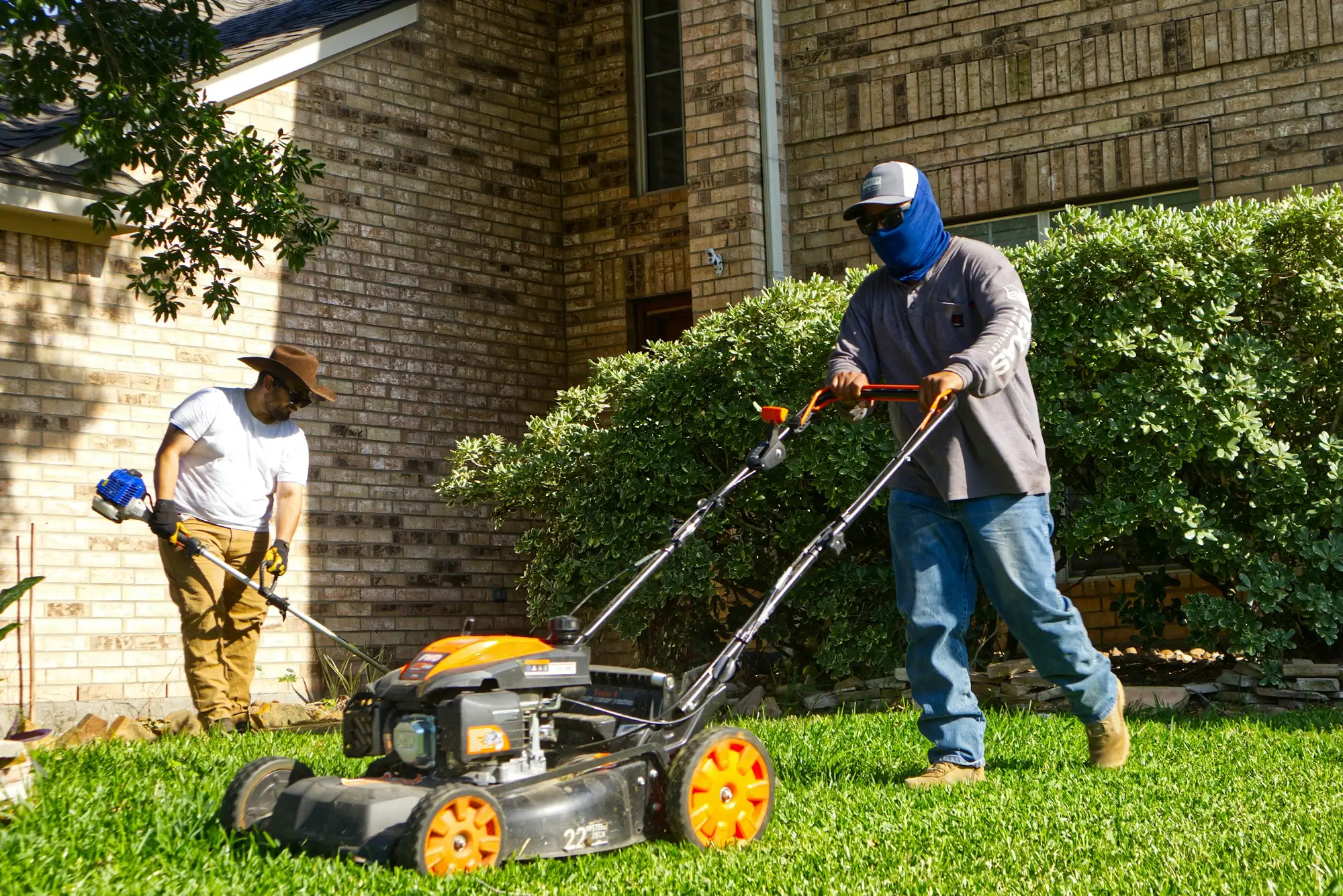 Two landscapers mowing and string trimming the front lawn of a brick house