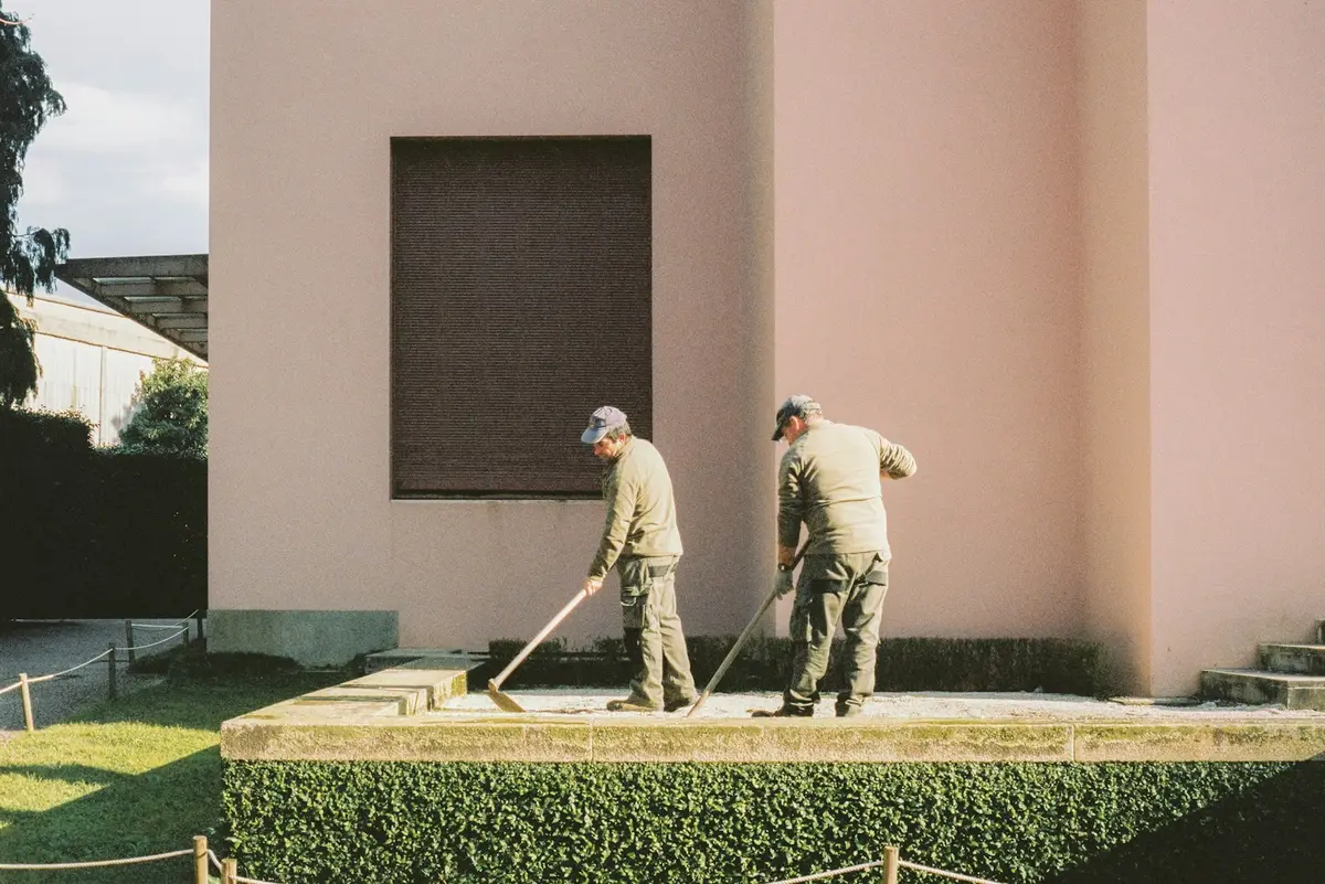 Landscaper in a yellow vest using a backpack leaf blower to clear fallen leaves from a lawn beside a large oak tree