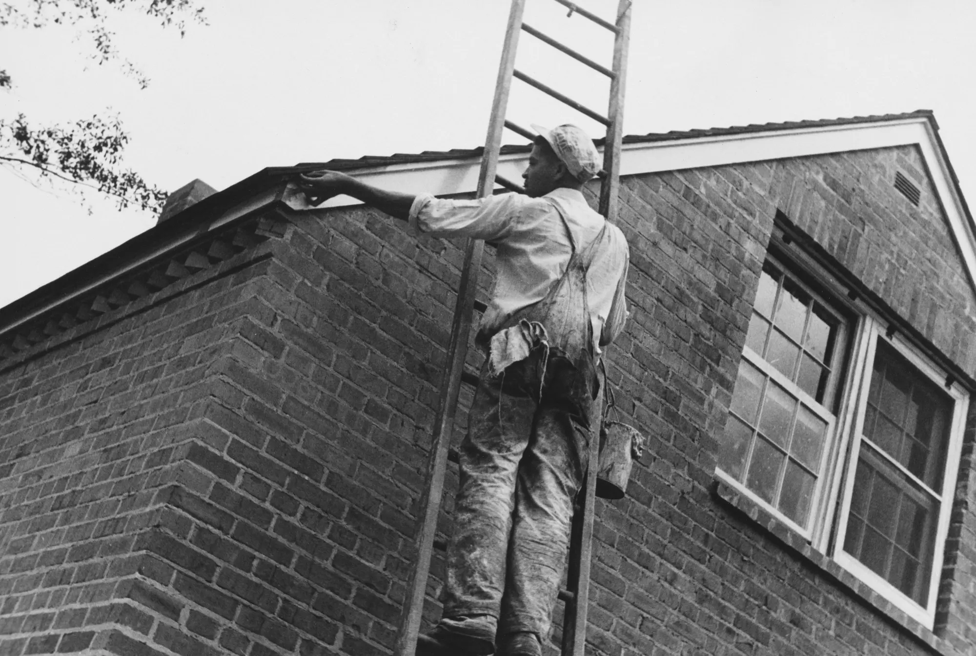 Painter on a wooden ladder painting the eave trim of a two-story brick house