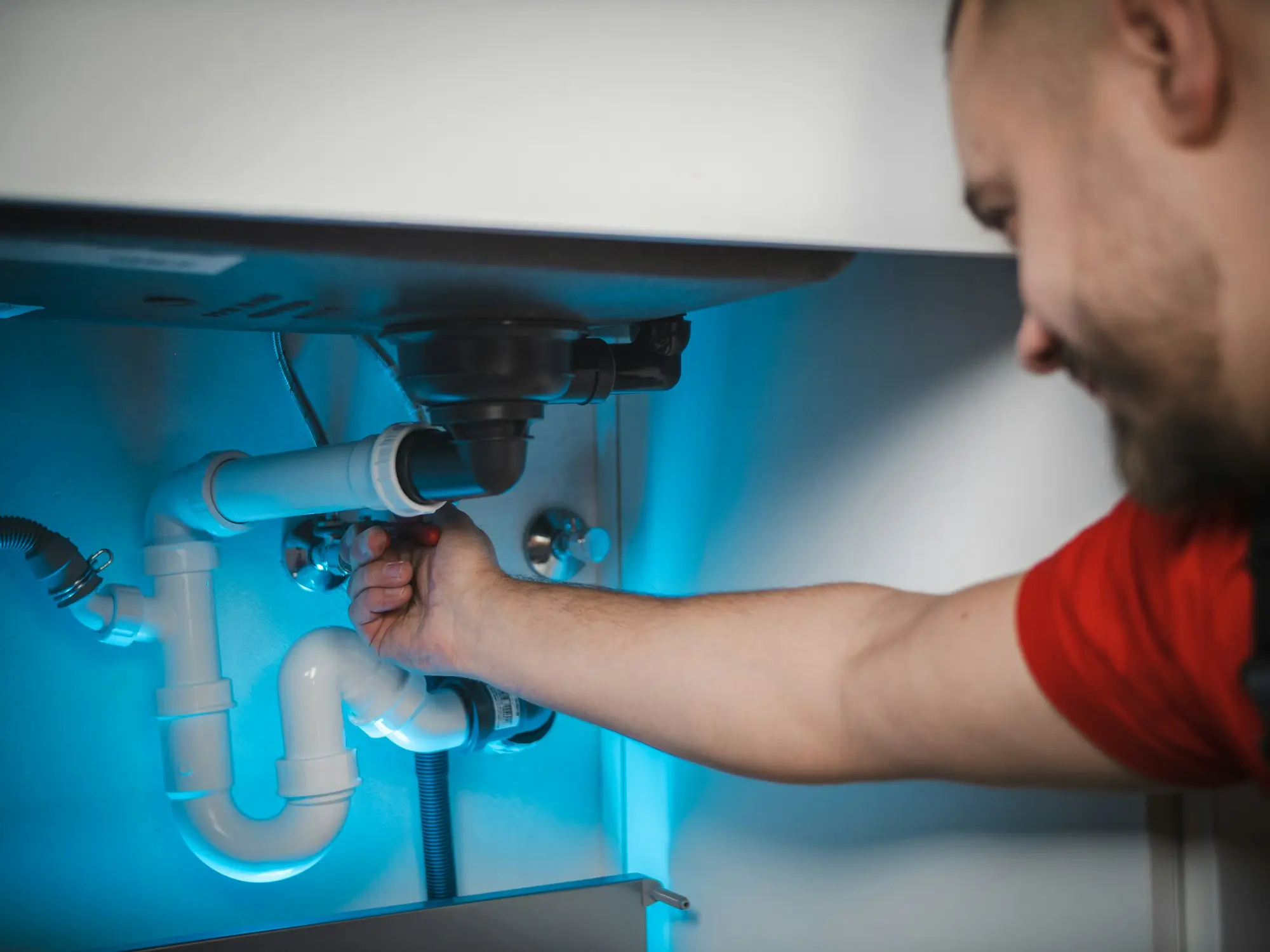 Plumber in a red shirt tightening a white PVC drain trap under a kitchen sink