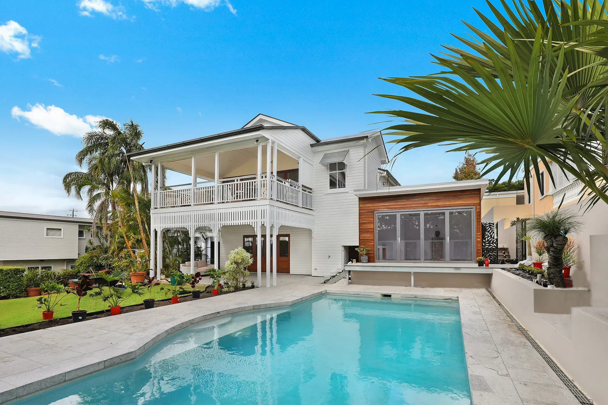 Backyard in-ground swimming pool with pale stone coping beside a white two-story house with palm trees